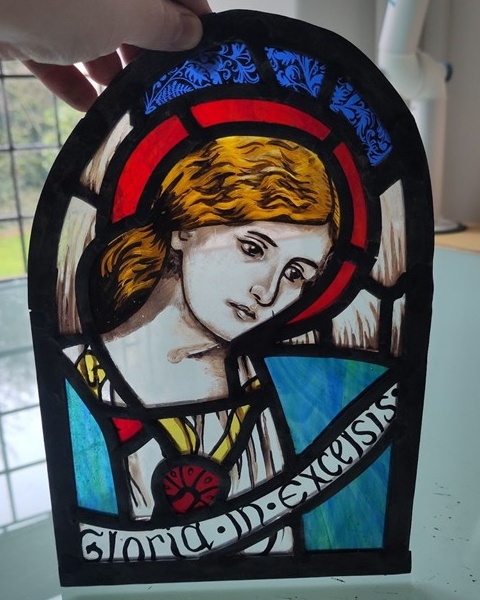 A stained glass panel depicting the bust of an angel with blue robes and a red halo. At the bottom is a banner which says 'Gloria in excelsius'